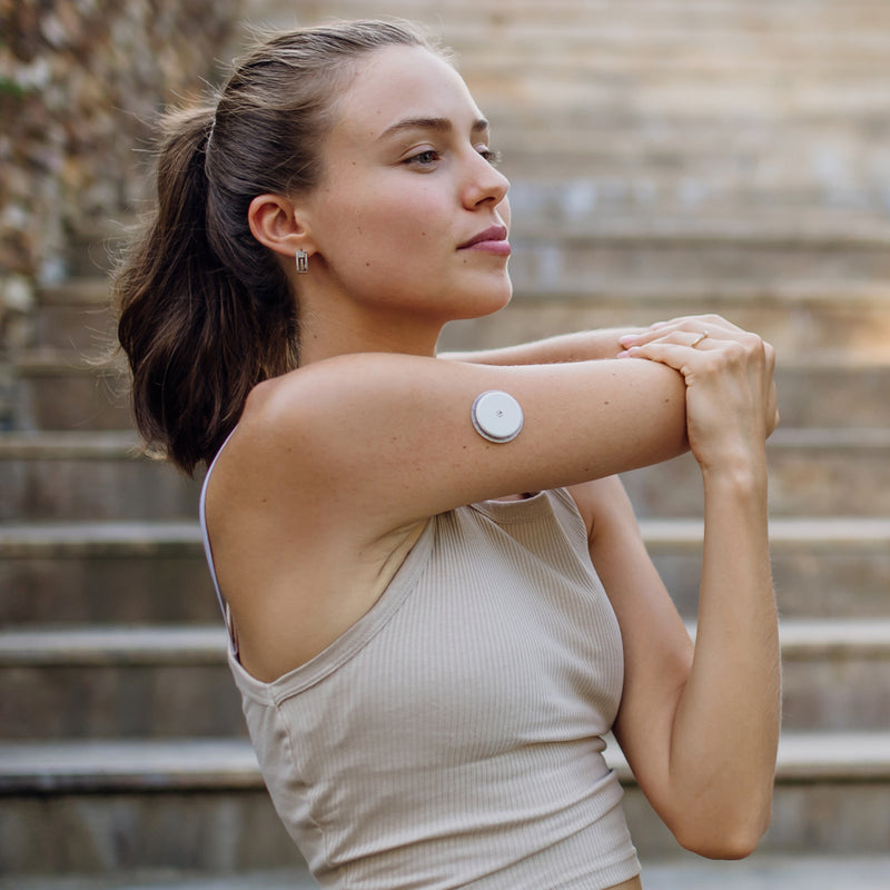 Woman with a glucose monitoring patch on her arm standing outdoors on steps
