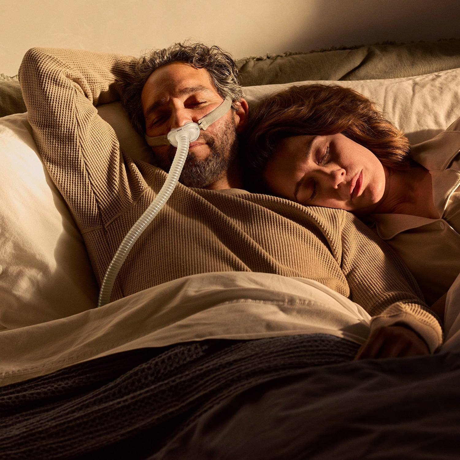 A man and a woman sleeping in bed under warm bedside lighting. The man wears a beige ribbed blanket and has a nasal CPAP Mask visible, while the woman rests beside him in a soft beige top.