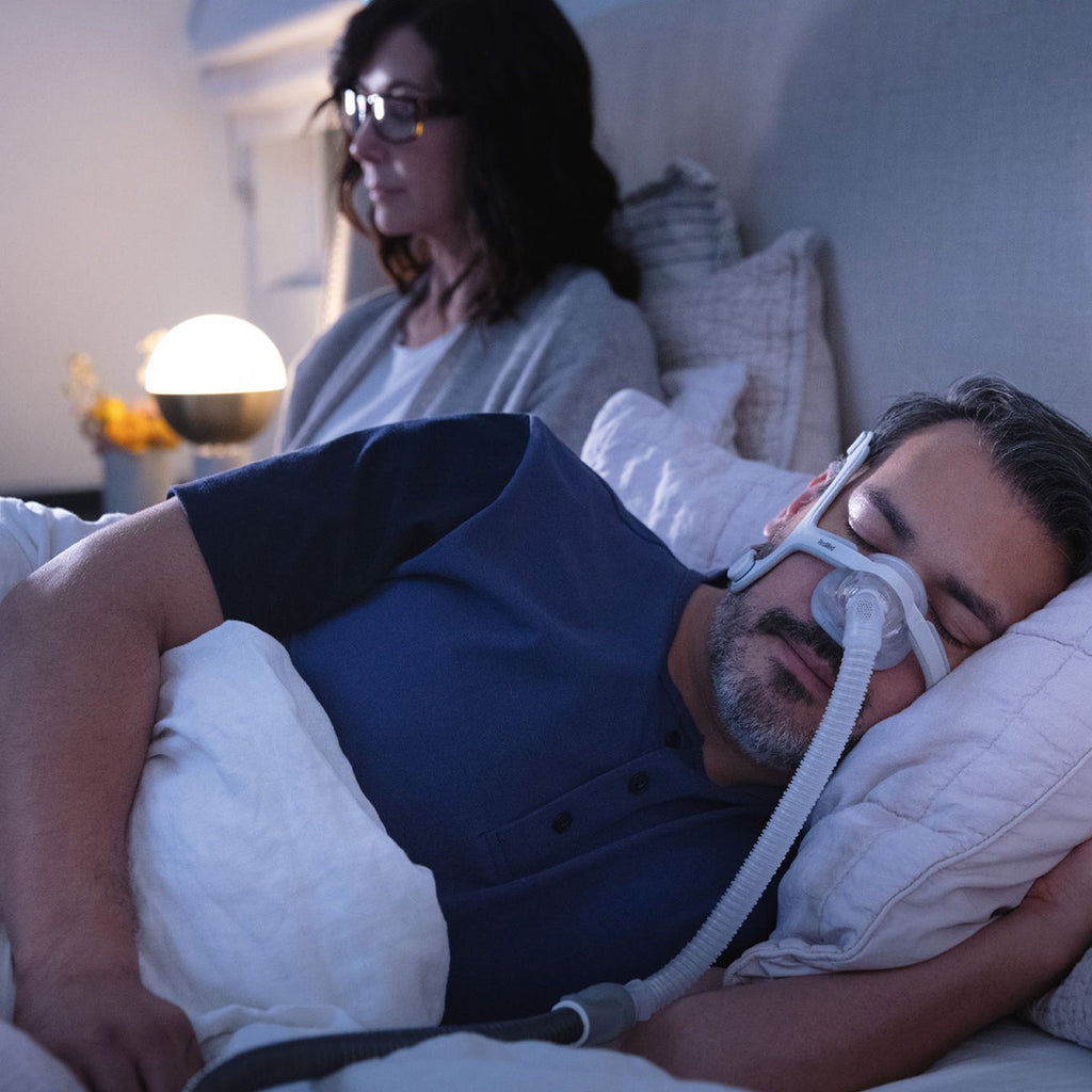 Man using a CPAP machine while a woman watches from behind.