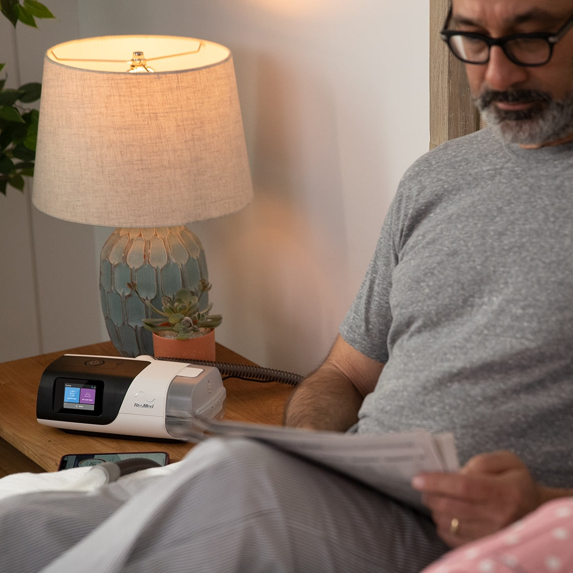 Man reading a newspaper next to a lamp and plant in a cozy room with a CPAP device next to the bed.