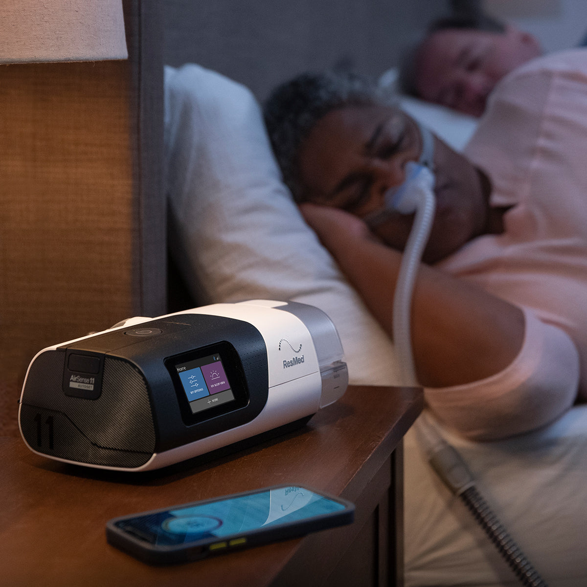 Person using a ResMed CPAP machine in bed with a smartphone on a nightstand.