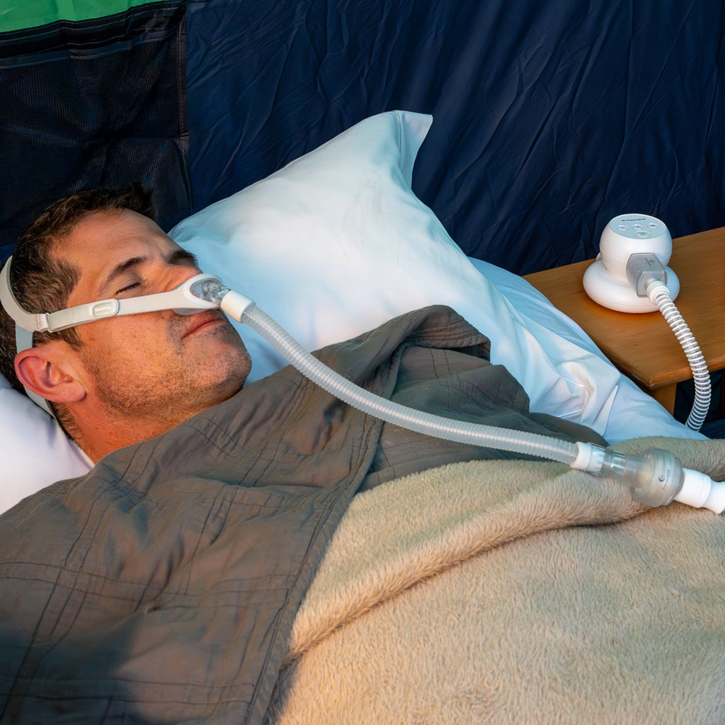 Man using a tiny CPAP machine in bed with a blue blanket and white pillow.