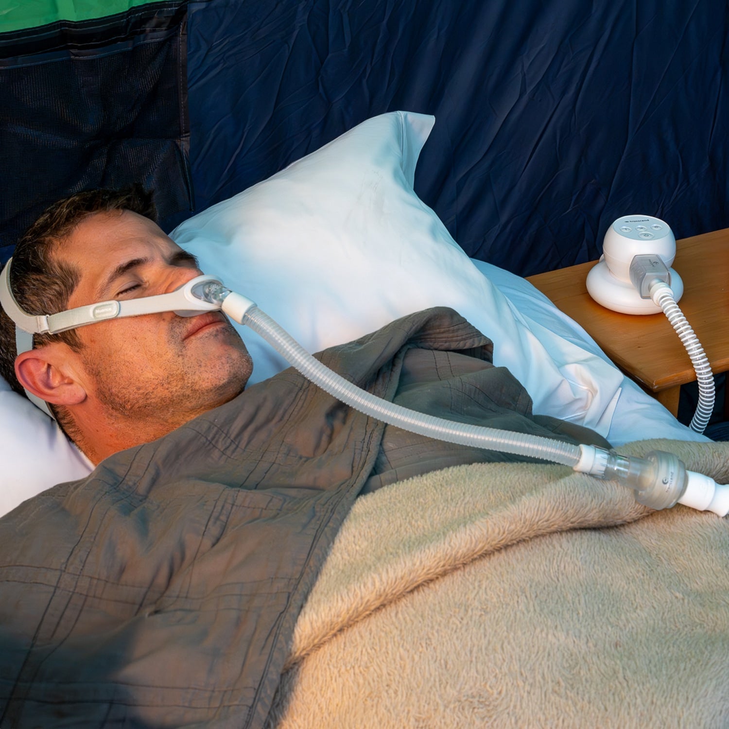Man using a tiny CPAP machine in bed with a blue blanket and white pillow.