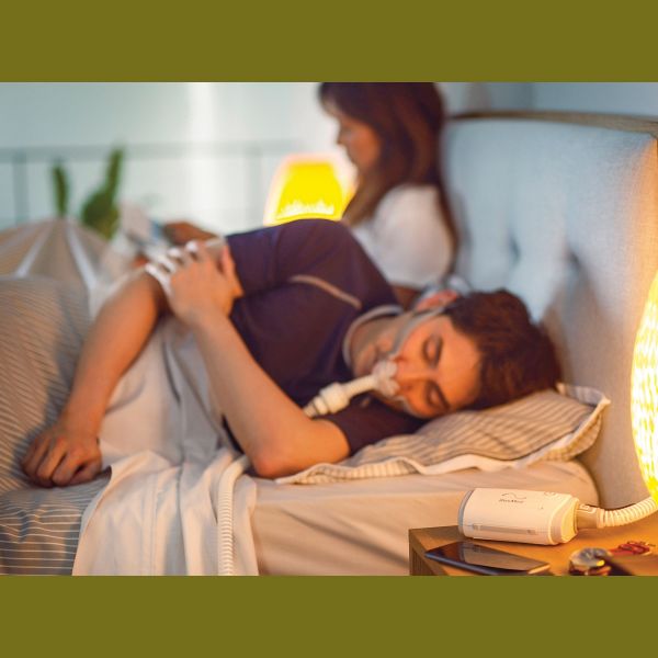 Man in bed with a CPAP mask on with a CPAP machine next to him and a women behind him reading